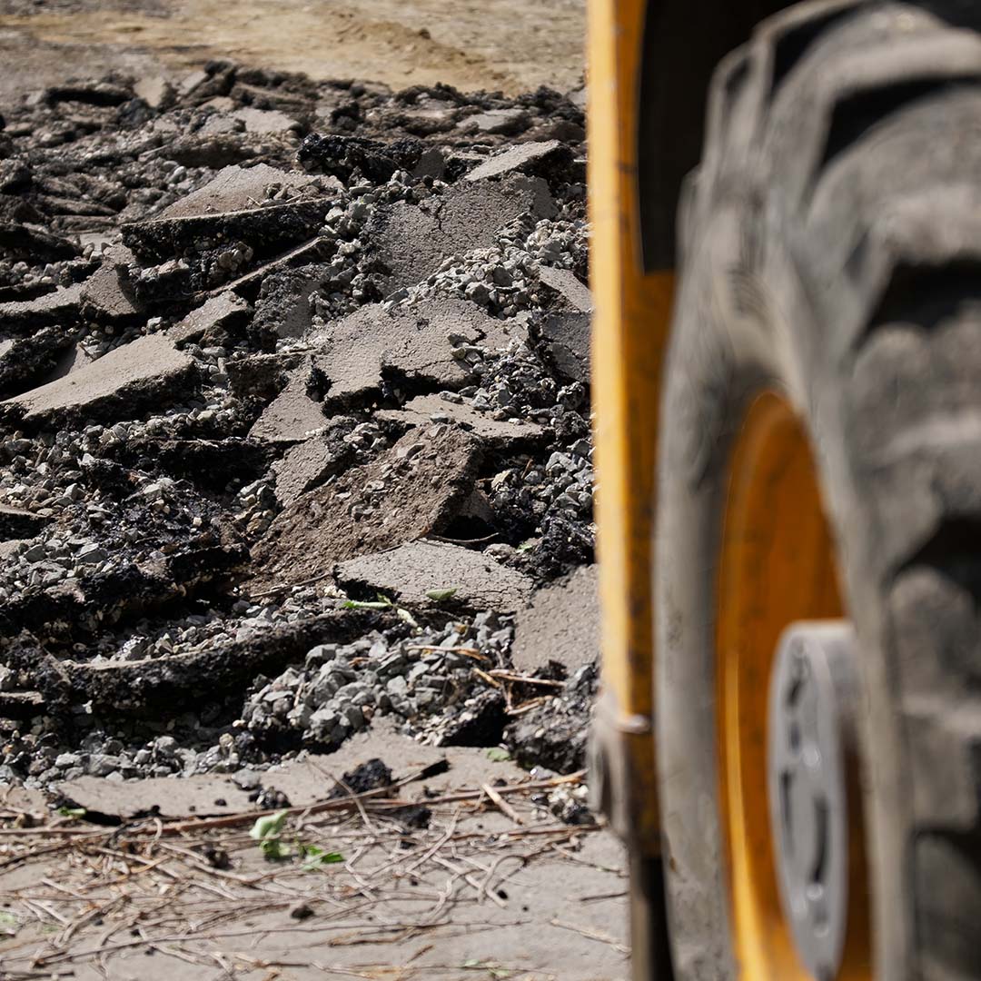 Broken asphalt on a city street next to the wheel of a heavy construction vehicle.